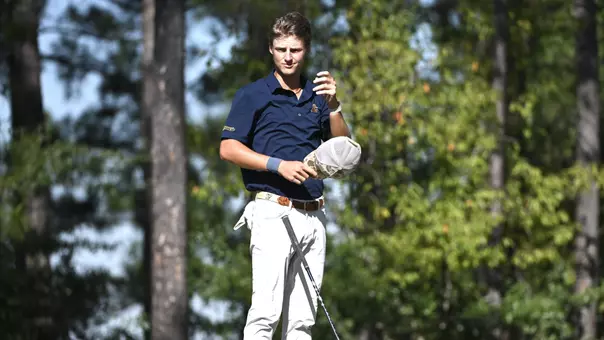 Evan Rogers musses his own hair with his hat in his hand at waist level awaiting his turn on the tee.