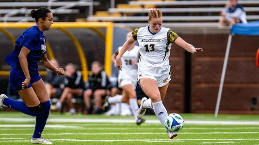 A Chattanooga soccer player wearing a white jersey with number 13 dribbles the ball up the field during a match while a defender in a purple uniform approaches from the left. The action takes place on a green turf field with team benches and stadium seating visible in the background.
