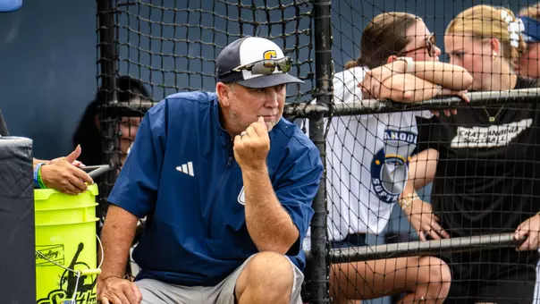 Chattanooga softball head coach watches from the dugout during a fall scrimmage at Frost Stadium as players look on from behind the fence.