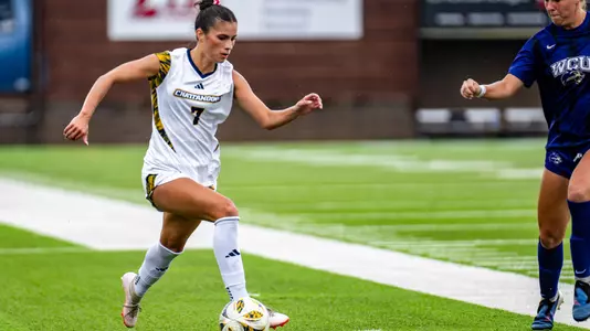 A soccer player in white is controlling the ball while a soccer player in dark is nearby.