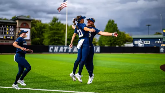Two softball players are on the field jumping in the air for a chest bump before a game.