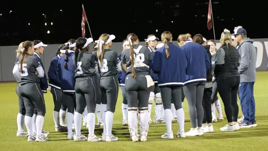 A softball team with coaches and players, gathers in a circle following a game.