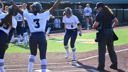 A softball player wearing number 6 steps on home plate during a home-run celebration as teammates raise their arms in excitement and an official observes the play near the batter’s box.
