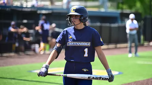 A softball player is holding a bat in front of her at waist level as she approaches the plate.