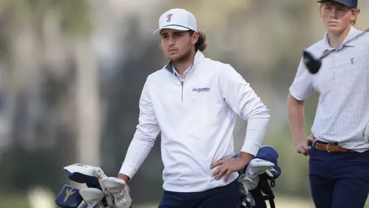 Ethan Whitaker looks down the fairway at his ball from the tee with a hand on his bag as he awaits playing partner's shot.