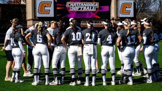 A softball team is gathered in a huddle in the outfield in front of a scoreboard that says Chattanooga Softball.