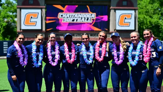Nine softball players wearing leis are standing in front of the scoreboard with their arms around each other.
