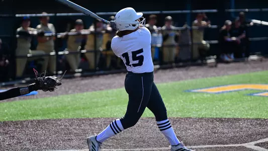 A softball player wearing white jersey and dark gray pants hit on her follow through after hitting a ball.