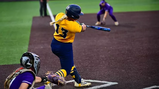 A softball player wearing a yellow top and blue pants hits the ball at the plate. The catcher is behind her.