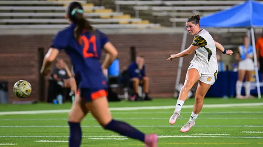 A soccer player in a white uniform kicks the ball downfield during a match while an opposing player in a navy uniform stands in the foreground.