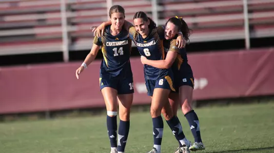 Three Chattanooga soccer players in navy and gold uniforms walk arm-in-arm on the field during a match. The player wearing No. 6 is between teammates wearing No. 14 and another teammate, with stadium bleachers visible in the background.