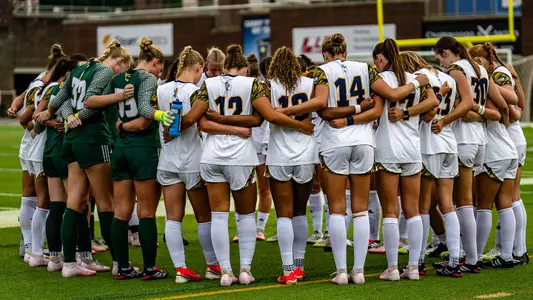 A soccer team in white and goalie green huddles together on the field before a game.