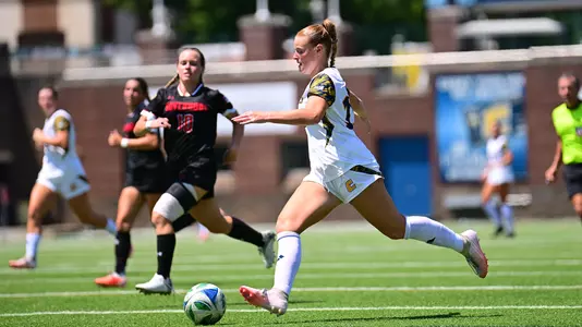 A women's soccer player in a white uniform dribbles the ball upfield during a match while two defenders in black uniforms pursue. The play takes place on a sunny afternoon with other players and an official visible in the background.