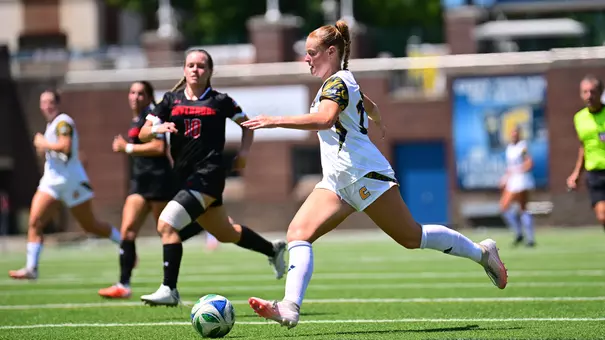 A women's soccer player in a white uniform dribbles the ball upfield during a match while two defenders in black uniforms pursue. The play takes place on a sunny afternoon with other players and an official visible in the background.