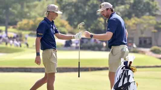 Evan Rogers with iron in hand and Coach Blaine Woodruff fist bump after a solid approach to the green in the background.