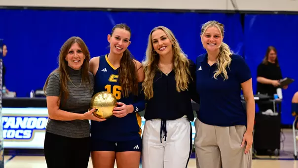 A volleyball player is receiving a gold ball from her head coach and assistant coaches after the game for her 1,000th career assist.