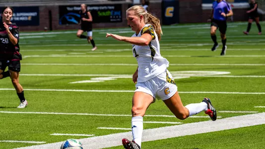 A soccer player in white is running down the field dribbling a ball with her feet.