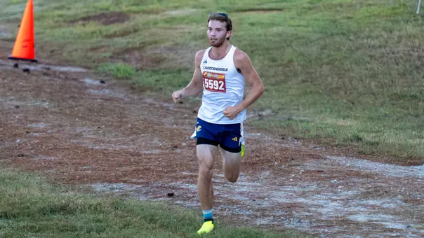A man is running in a college cross country race on a grassy course.