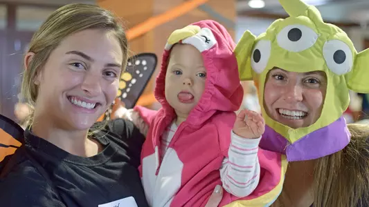 Badgers Give Back Spooktacular event - Two UW student-athletes in Halloween costumes hold a small girl from Wisconsin Upside Down and smile.