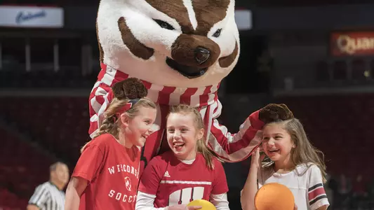 Bucky Badger giving toy basketballs to three young girls at women's basketball game