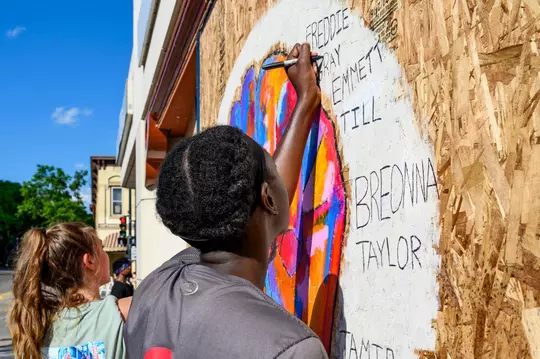 Students painting murals on State Street 2020 over plywood from destroyed windows due to racial justice protests. The students write the names of victims of racial injustice murders such as Breonna Taylor, Emmett Till and George Floyd
