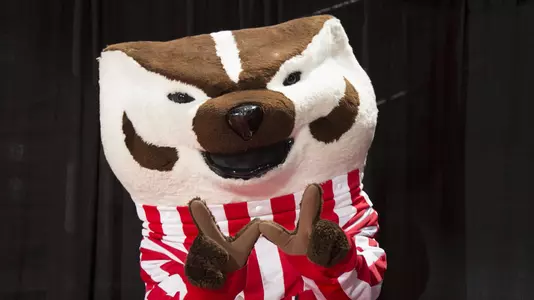 Bucky Badger holds his paws in W for Wisconsin. Bucky is standing against a black background.