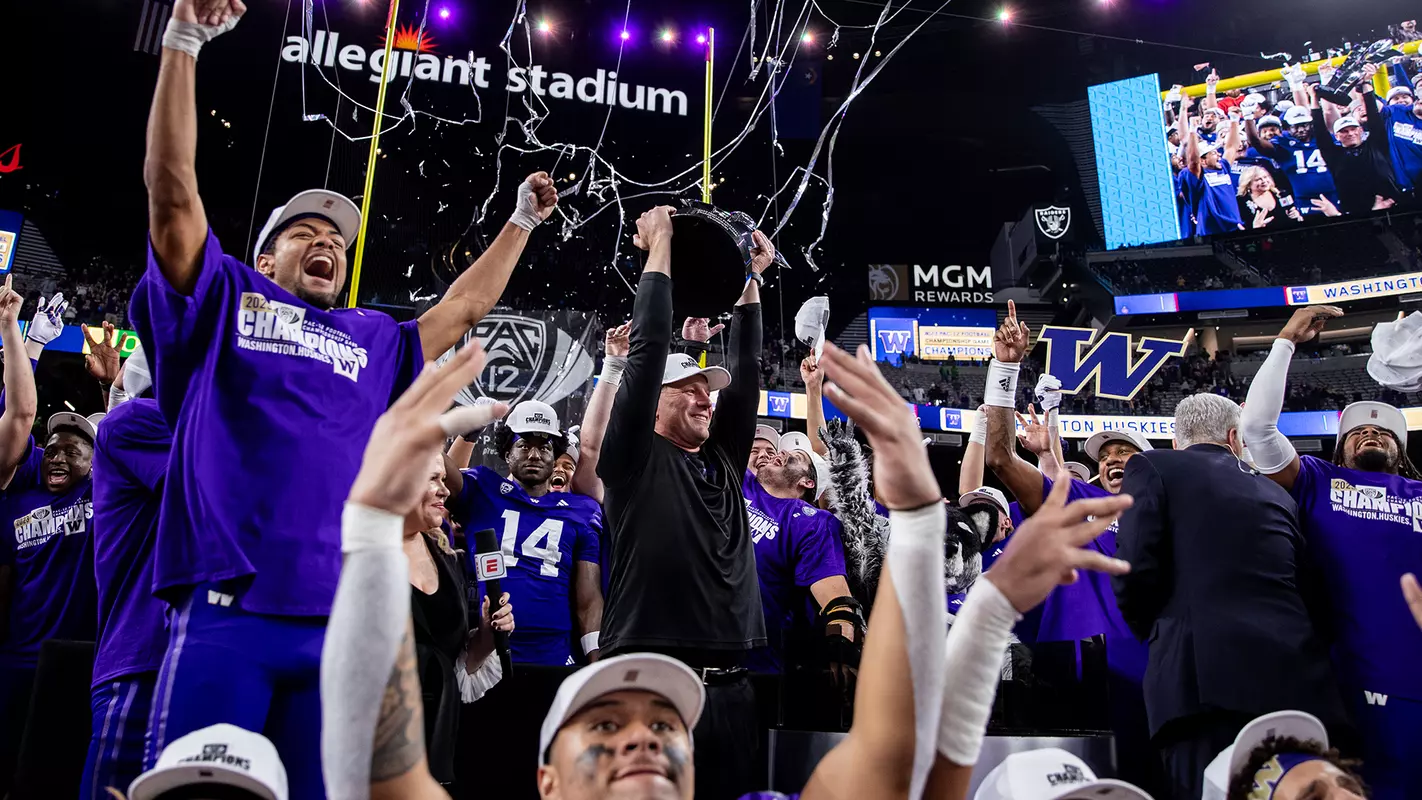 Coach DeBoer holding Pac-12 championship trophy with players celebrating at Allegiant Stadium