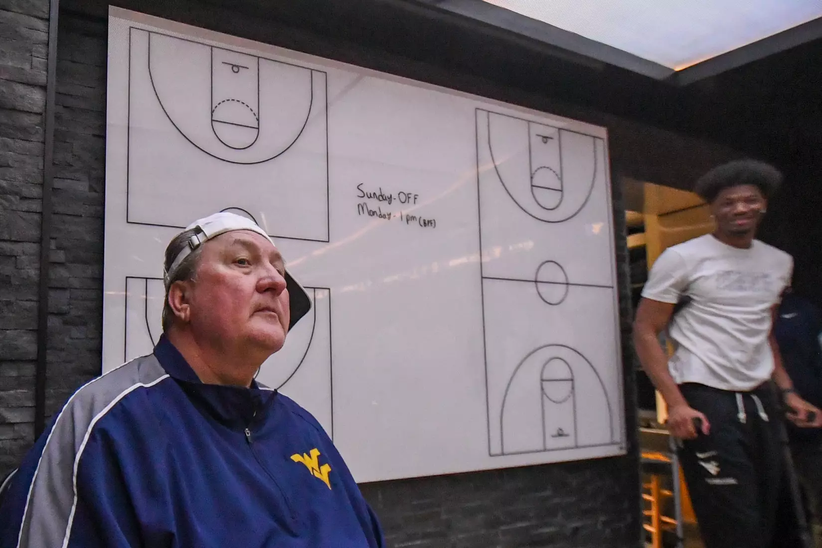 Bob Huggins gets a sideways-turned hat from his team in the locker room as Mohamed Wague watches.