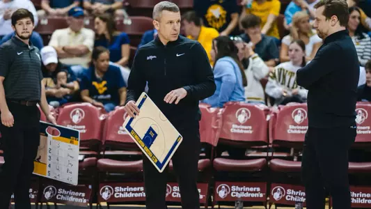 Ross Hodge stands on the sideline holding a clipboard during a WVU men's basketball game.