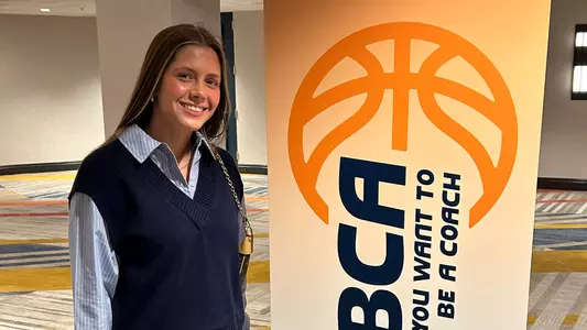 Casey Santoro stands to the left of a vertical sign for the WBCA So You Want To Be A Coach program