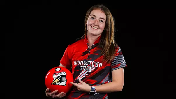Elizabeth Teuber holds and points to a red bowling ball with YSU's Pete the Penguin logo in a studio setting.
